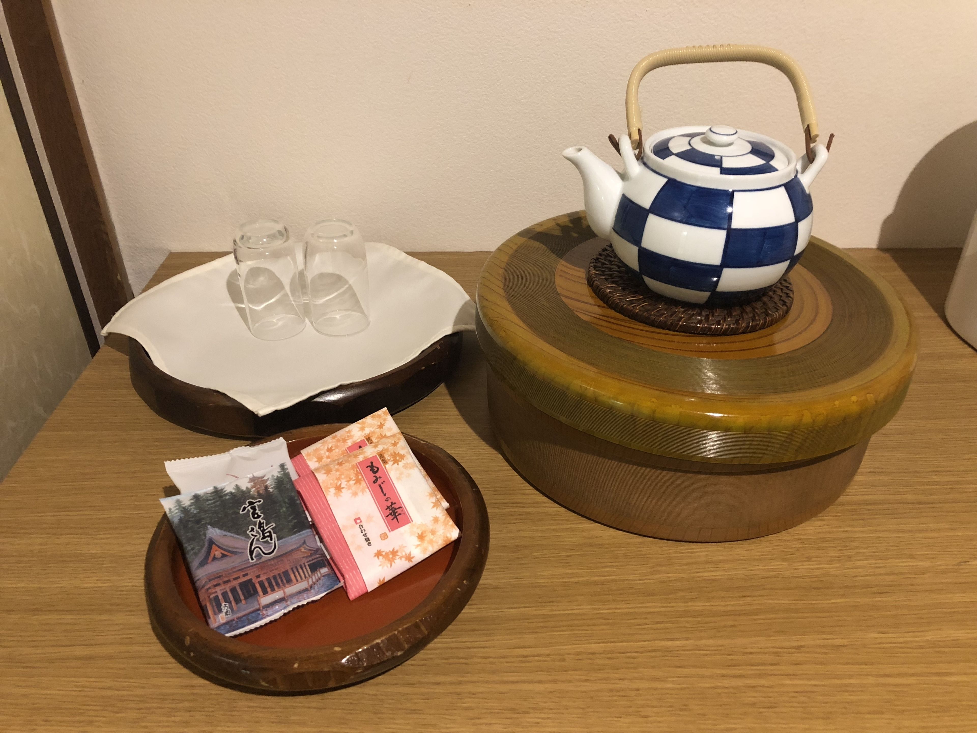Una mesa de madera con una tetera azul y blanca a cuadros, dos vasos transparentes, una bandeja blanca con un paño y una bandejita con una bolsita de té y una imagen.