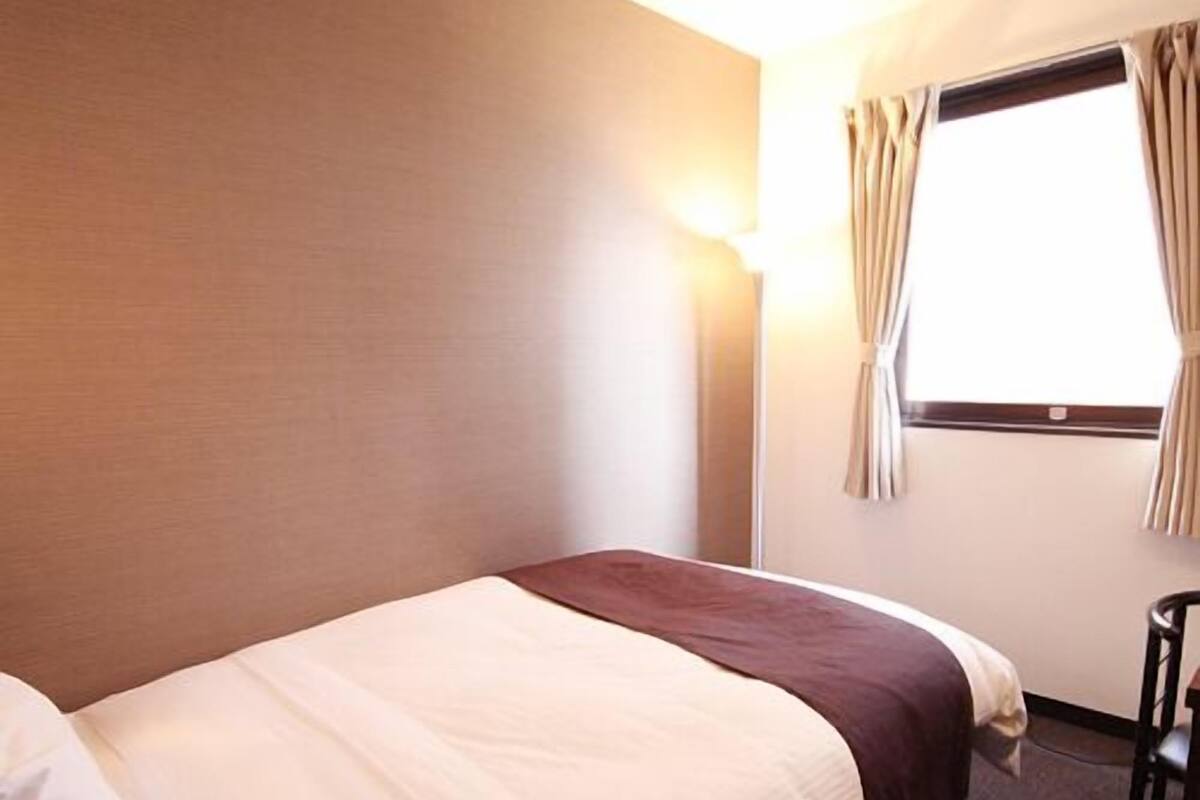 A single bed with white and brown bedding, a nightstand, a lamp, and a window with curtains.