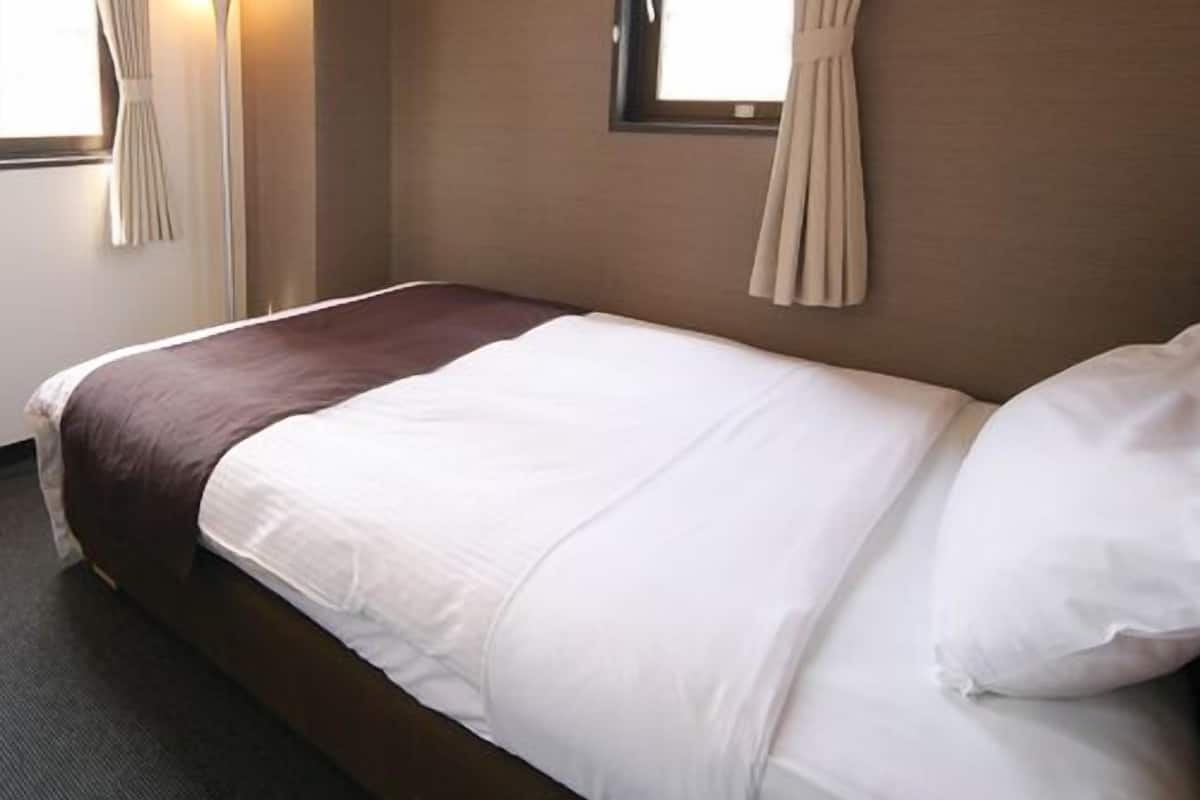 A hotel room with a bed, curtains, a window, and a wall lamp.