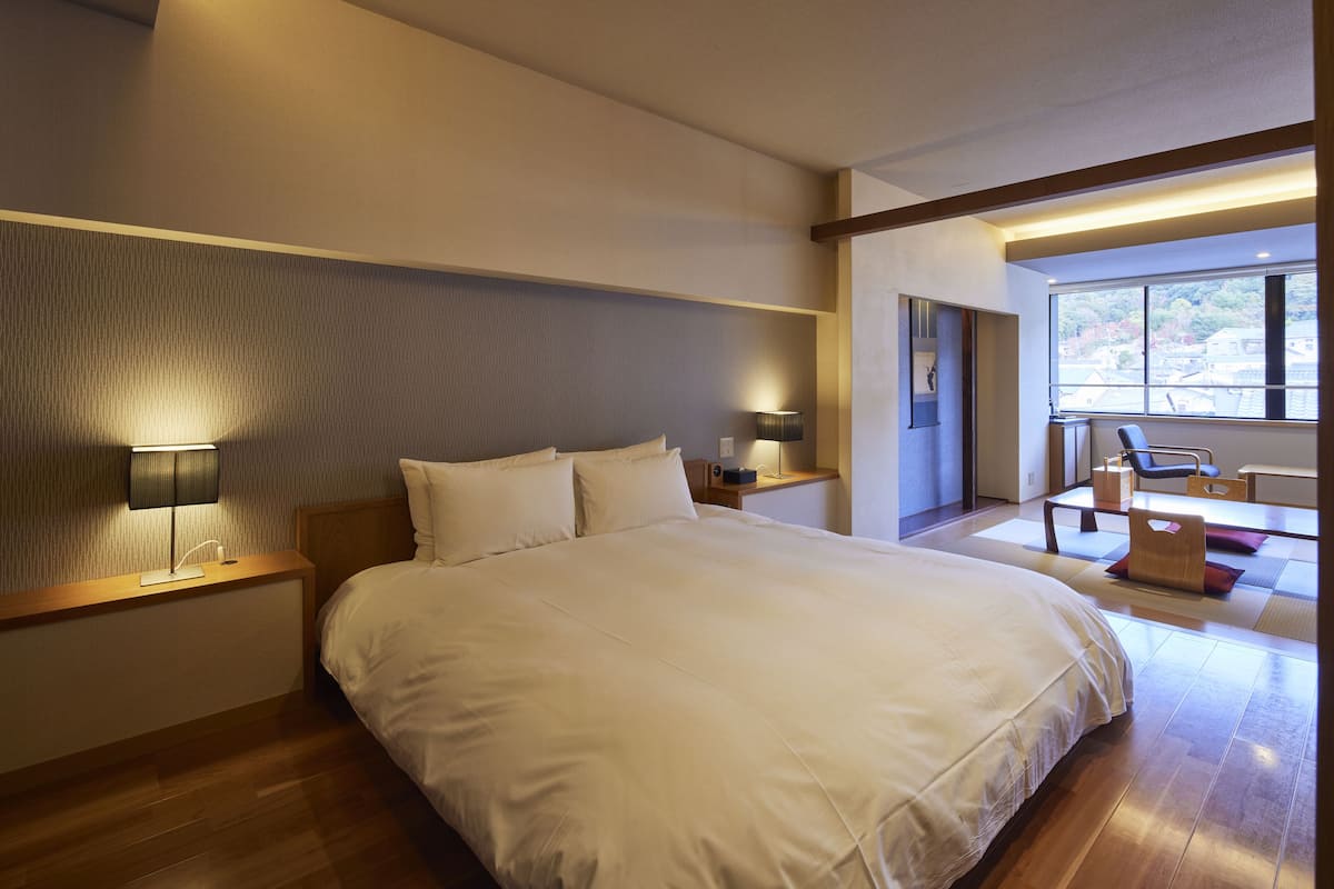 A modern hotel room with a large bed, bedside lamps, a small table, and a view of the outdoors through a large window.