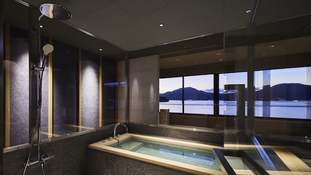 A modern bathroom with a glass-enclosed jacuzzi, a shower, and a view of a lake and mountains. - Itsukushima Iroha  (Hatsukaichi)