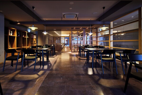 Un interior de restaurante moderno con mesas y sillas altas, estanterías de madera y ventanas amplias que dejan entrar luz natural.