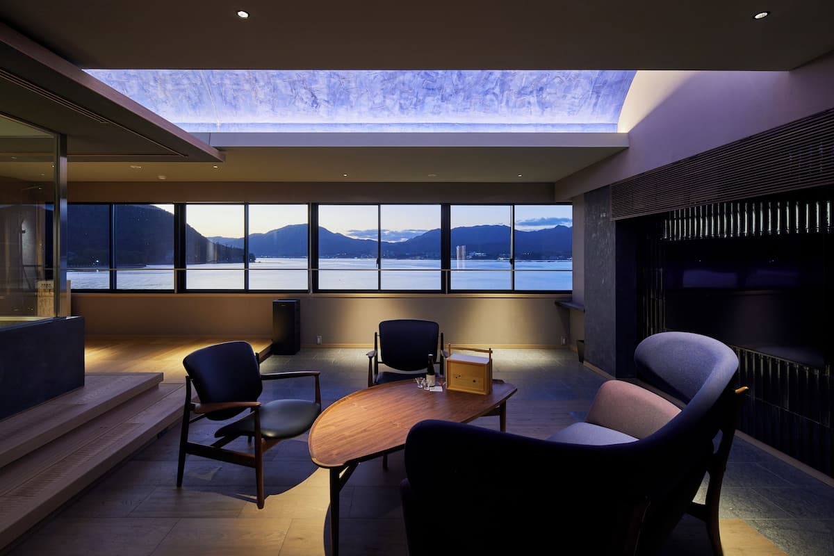 A modern interior with a large window overlooking a lake and mountains, a wooden table, and several chairs.