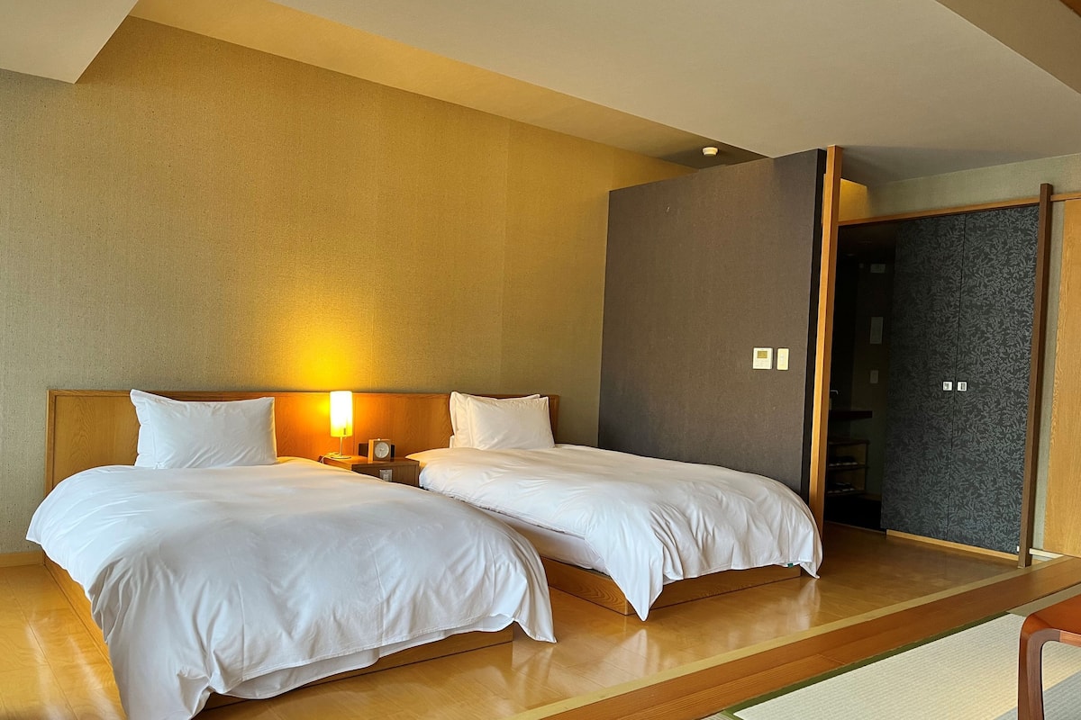 A hotel room with two beds, wooden headboards, a nightstand with a lamp, and a wardrobe with a patterned door.