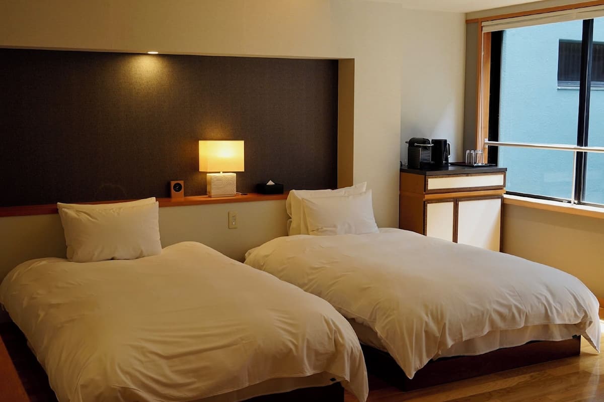 A hotel room with two beds, a bedside table with a lamp, a coffee maker, and a window with a view.