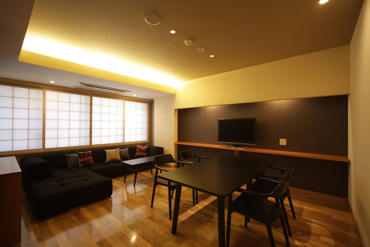 A modern living room with a black sofa, a dining table with chairs, and a flat-screen TV mounted on the wall.