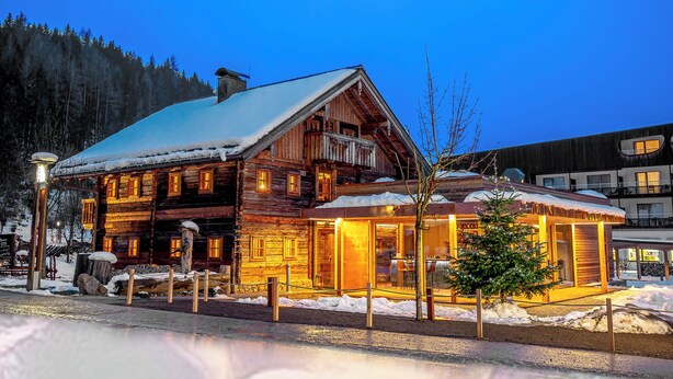 Eine Holzhütte mit schneebedecktem Dach und beleuchtetem Innenraum, umgeben von Schnee und Bäumen.