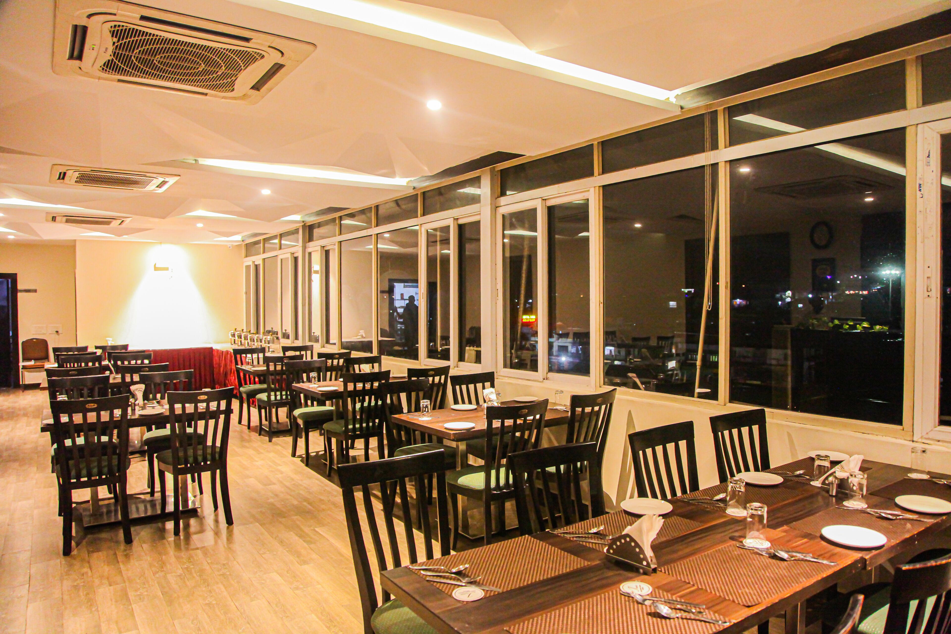 Restaurante com várias mesas e cadeiras, janelas amplas e vista da cidade à noite.