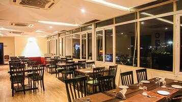 Restaurante com várias mesas e cadeiras, janelas amplas e vista da cidade à noite.
