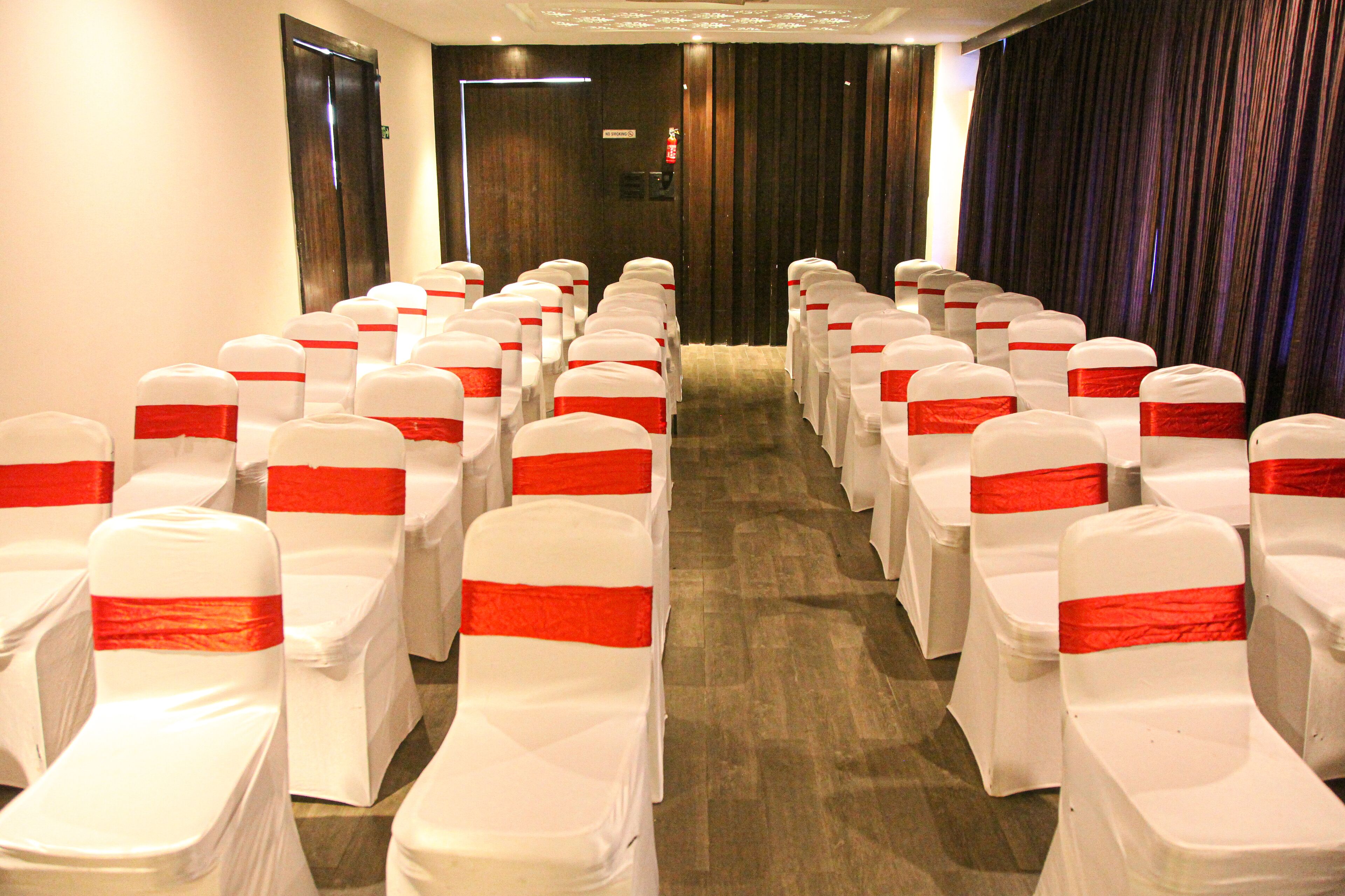 Un salón de conferencias con filas de sillas blancas adornadas con lazos rojos, suelo de madera y un telón oscuro de fondo.