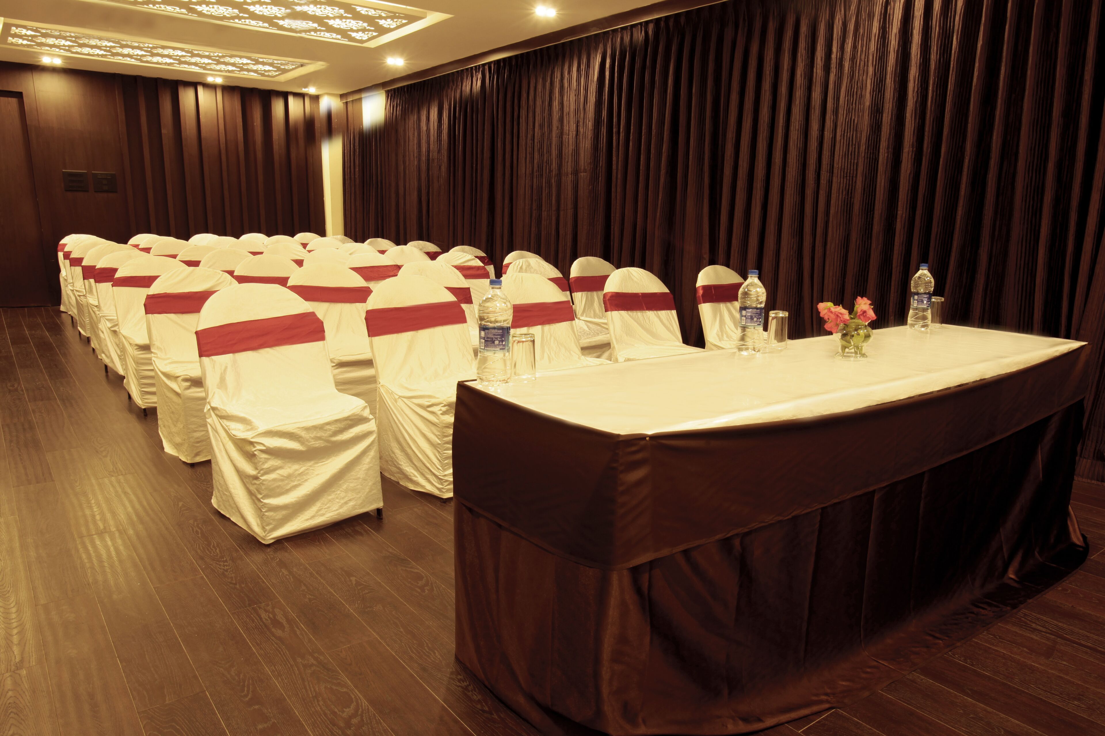 Une salle de conférence avec des rangées de chaises recouvertes de nappes blanches et ornées de rubans rouges, une longue table avec des bouteilles d’eau et des verres, et un rideau sombre en arrière-plan.