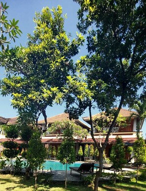 Zona de piscina con sillones de descanso, rodeada de exuberante vegetación y un edificio con techo de tejas.