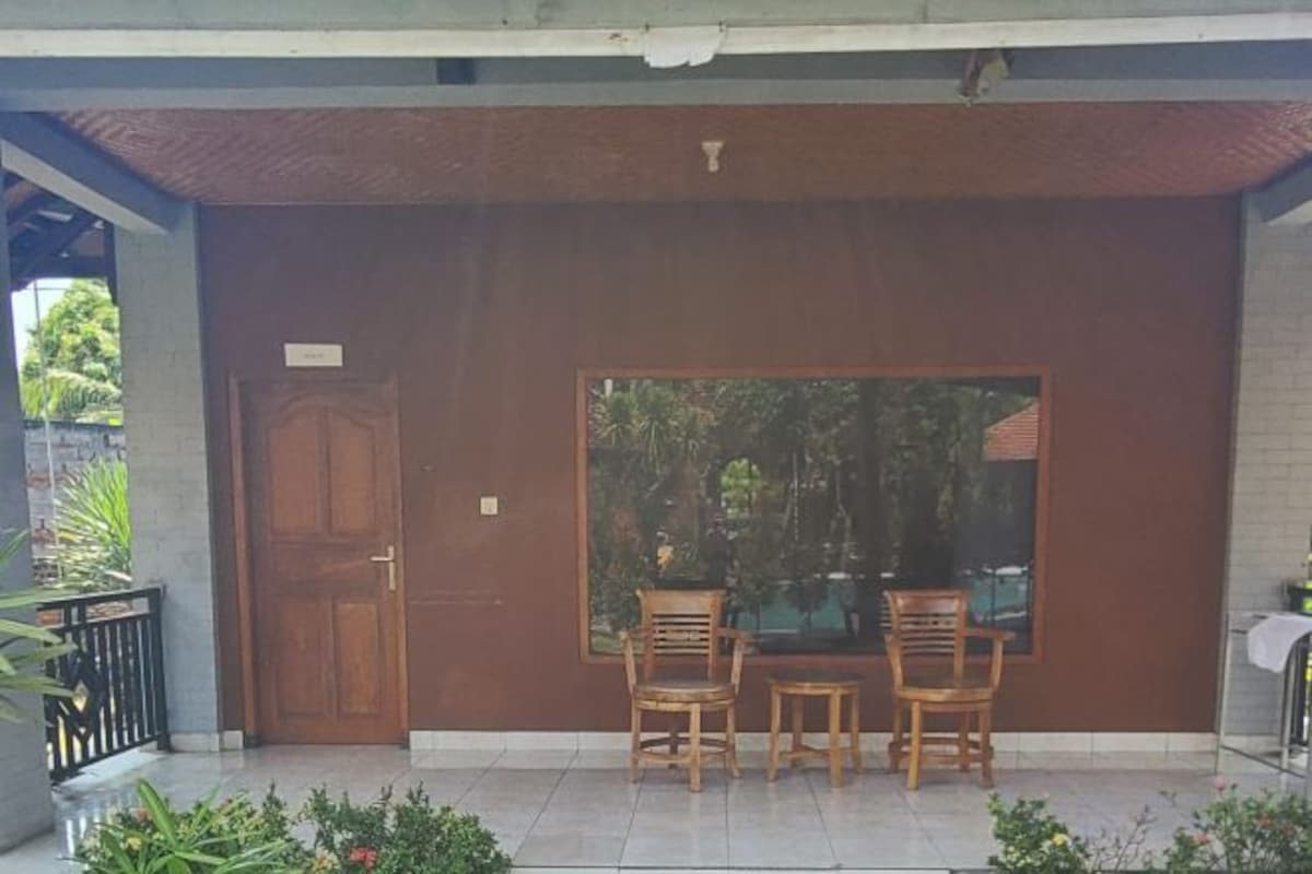 A covered outdoor seating area with two wooden chairs and a table, a wooden door, and a large window with a view of greenery.