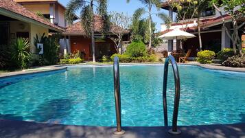 A clear swimming pool with metal ladders, surrounded by greenery and buildings with red-tiled roofs.