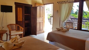 A room with two beds, a wooden chair, a TV, and a balcony with a view of greenery. - Hotel Mylovina (Buleleng)