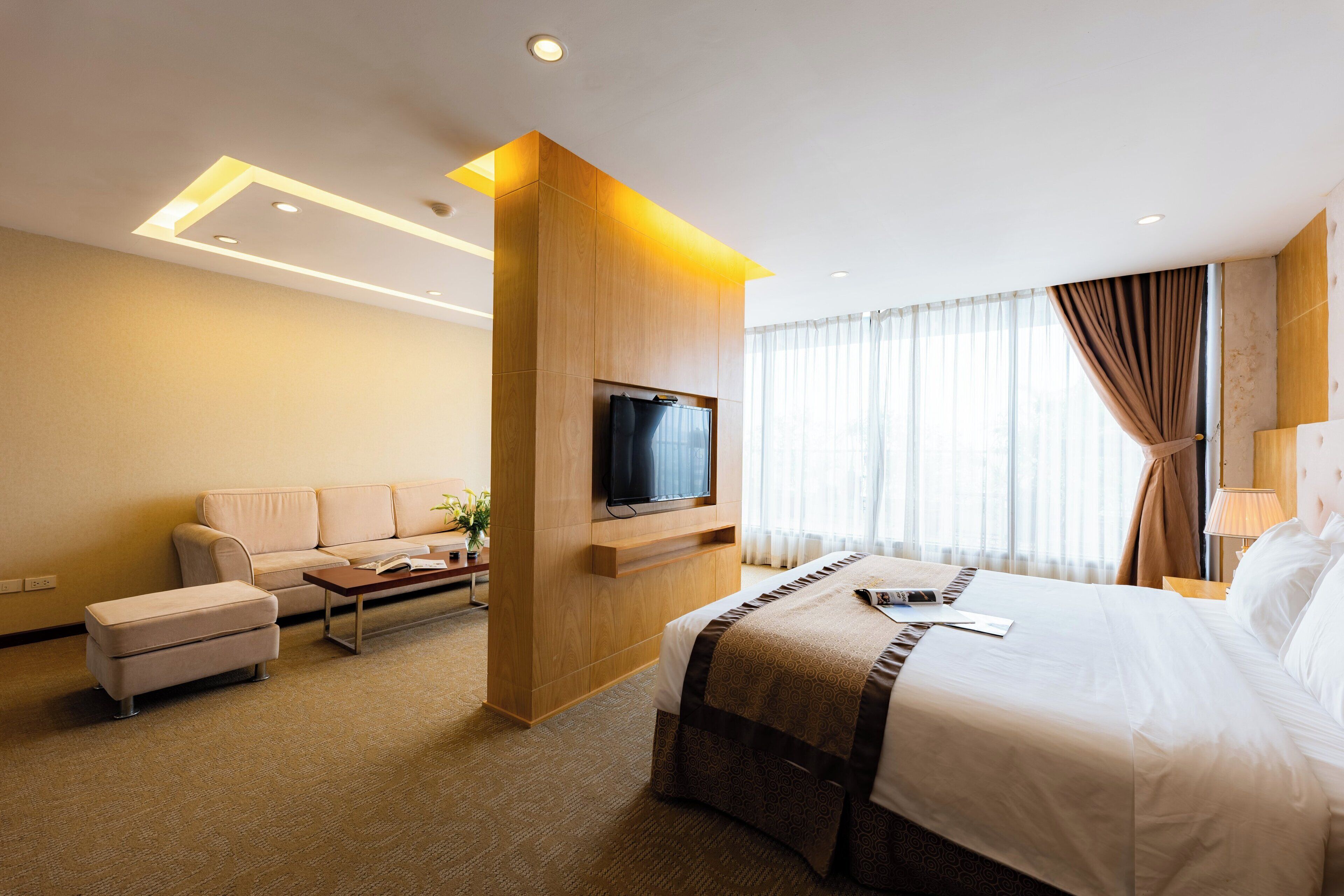 A modern hotel room with a large bed, a flat-screen TV mounted on a wooden panel, a sofa, and a small table.