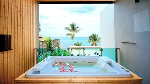 Un balcone in legno con una vasca idromassaggio piena d'acqua e petali di fiori, con vista su una spiaggia, palme e l'oceano.