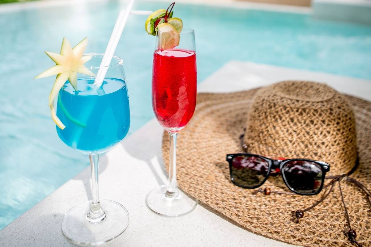 Two drinks on a poolside table with a straw, a straw hat, and sunglasses.