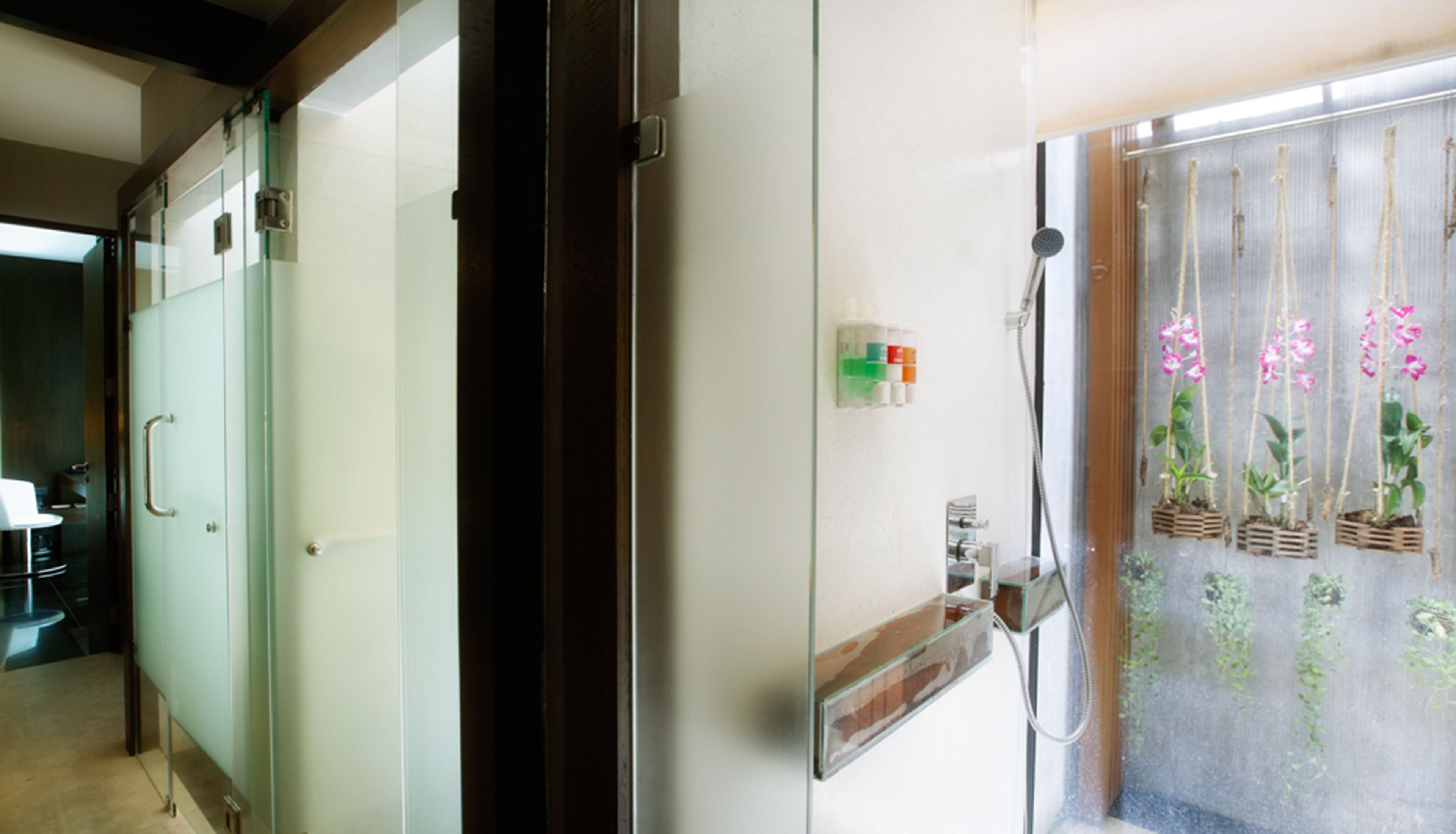 Quarto de hotel com porta deslizante para um banheiro, chuveiro com box de vidro e vista para orquídeas através do vidro.