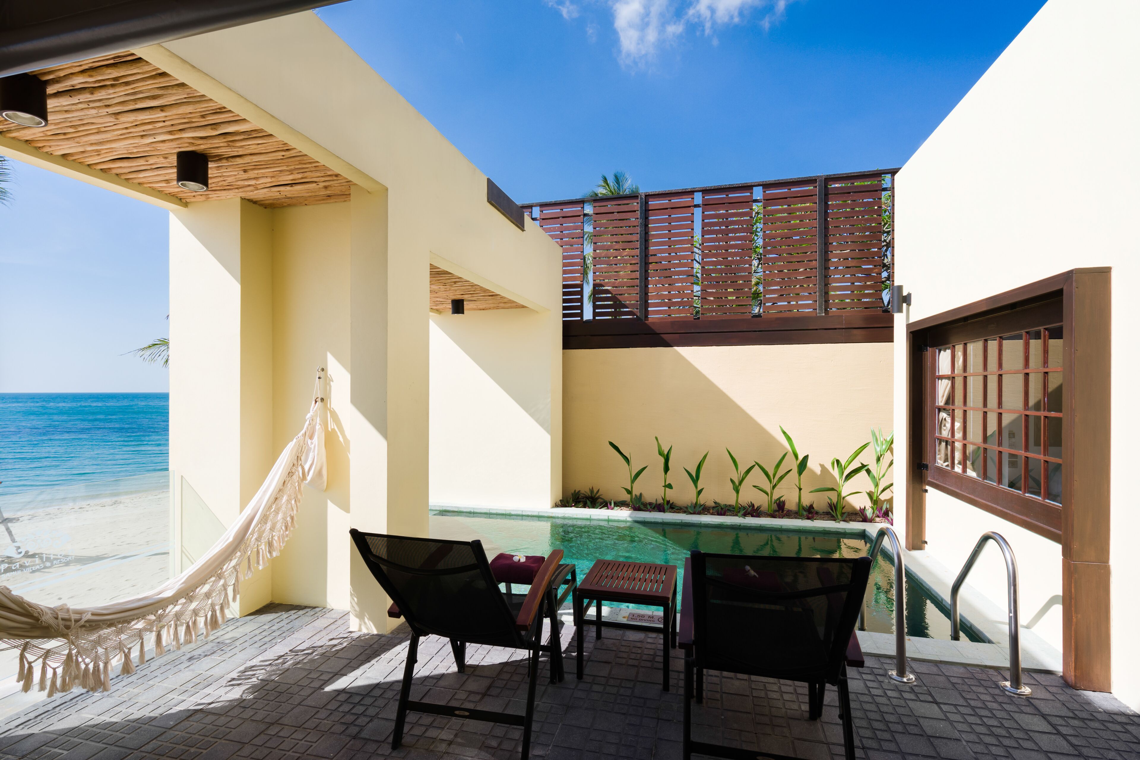 A poolside area with a hammock, chairs, and a table, overlooking a beach and a clear sky.