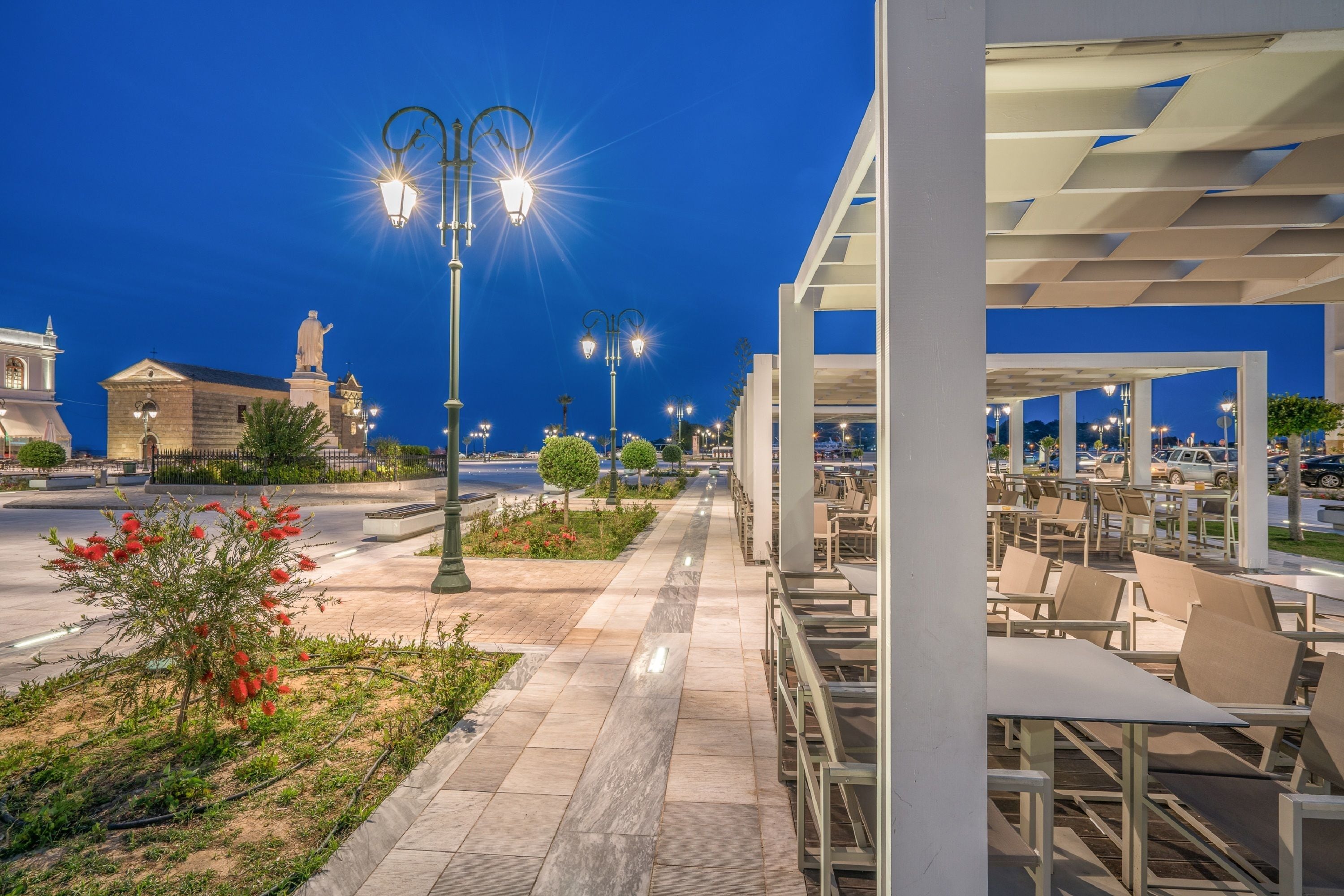 Een buitenzitplaats met tafels en stoelen, een pad en een gebouw met een standbeeld op de achtergrond.