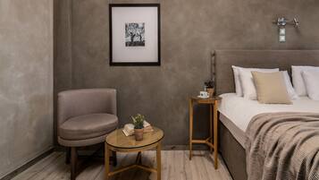 Una habitación con cama, una silla, una mesita con una planta y un cuadro en la pared.