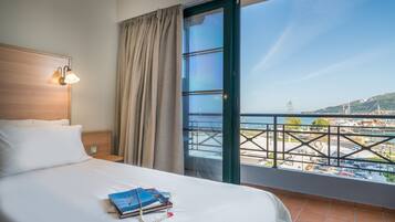 Una camera d'albergo con un letto, un comodino, una lampada, una finestra che si affaccia su un porto turistico e degli edifici, e una tenda.