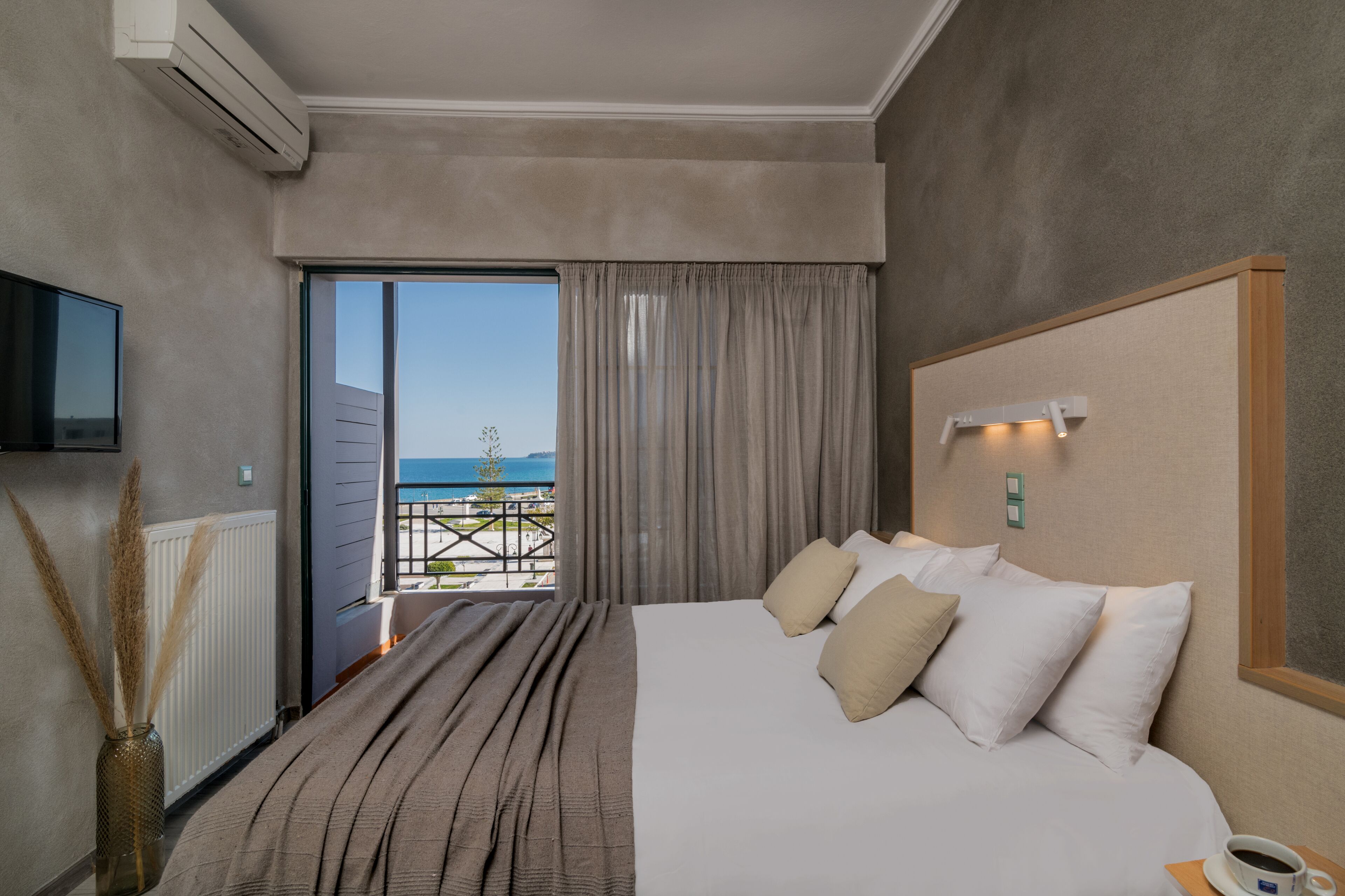 Une chambre d’hôtel avec un grand lit, un téléviseur fixé au mur, une vue sur la mer depuis un balcon et un appareil de climatisation.