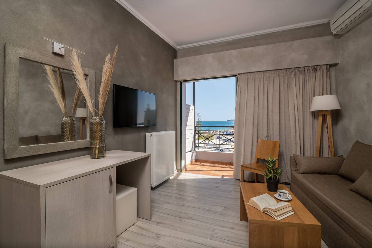 A modern living room with a flat-screen TV, a wooden coffee table with books and a cup, a sofa, and a view of the sea through large windows.