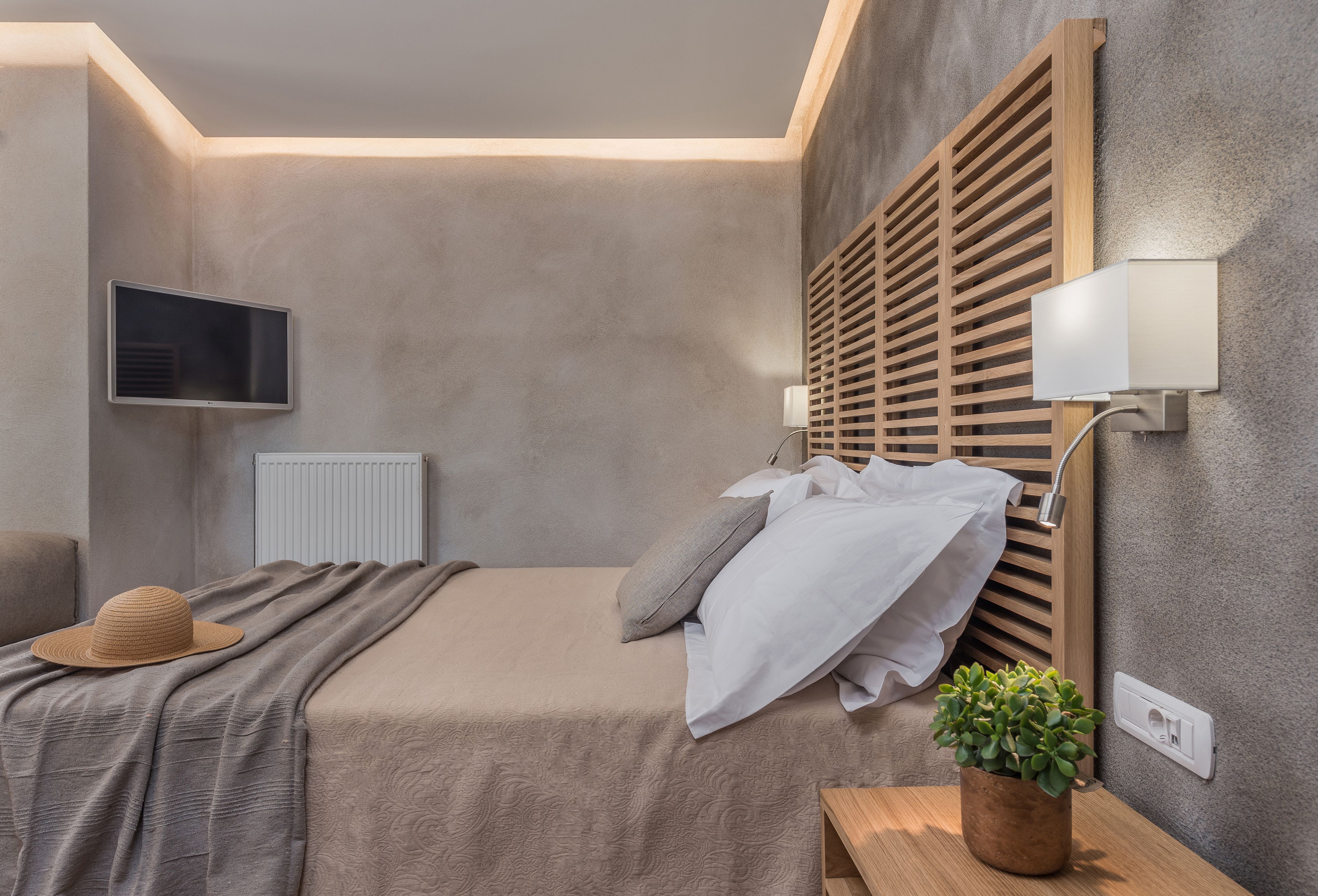 Un dormitorio con cama, televisión montada en la pared, cabecero de madera y mesita de noche con una planta.