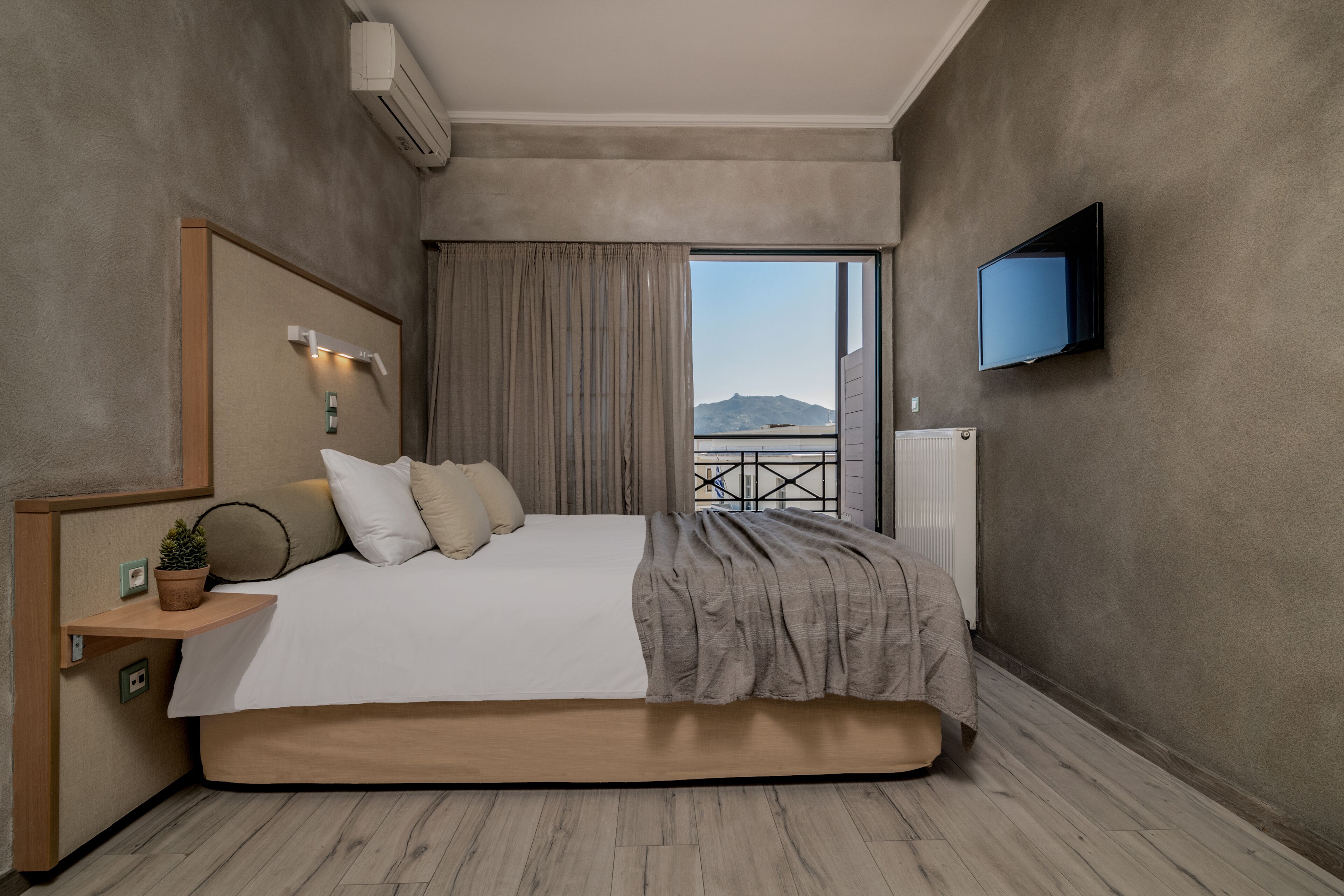 A modern bedroom with a large bed, a mounted TV, and a view of a balcony with a mountain in the background.