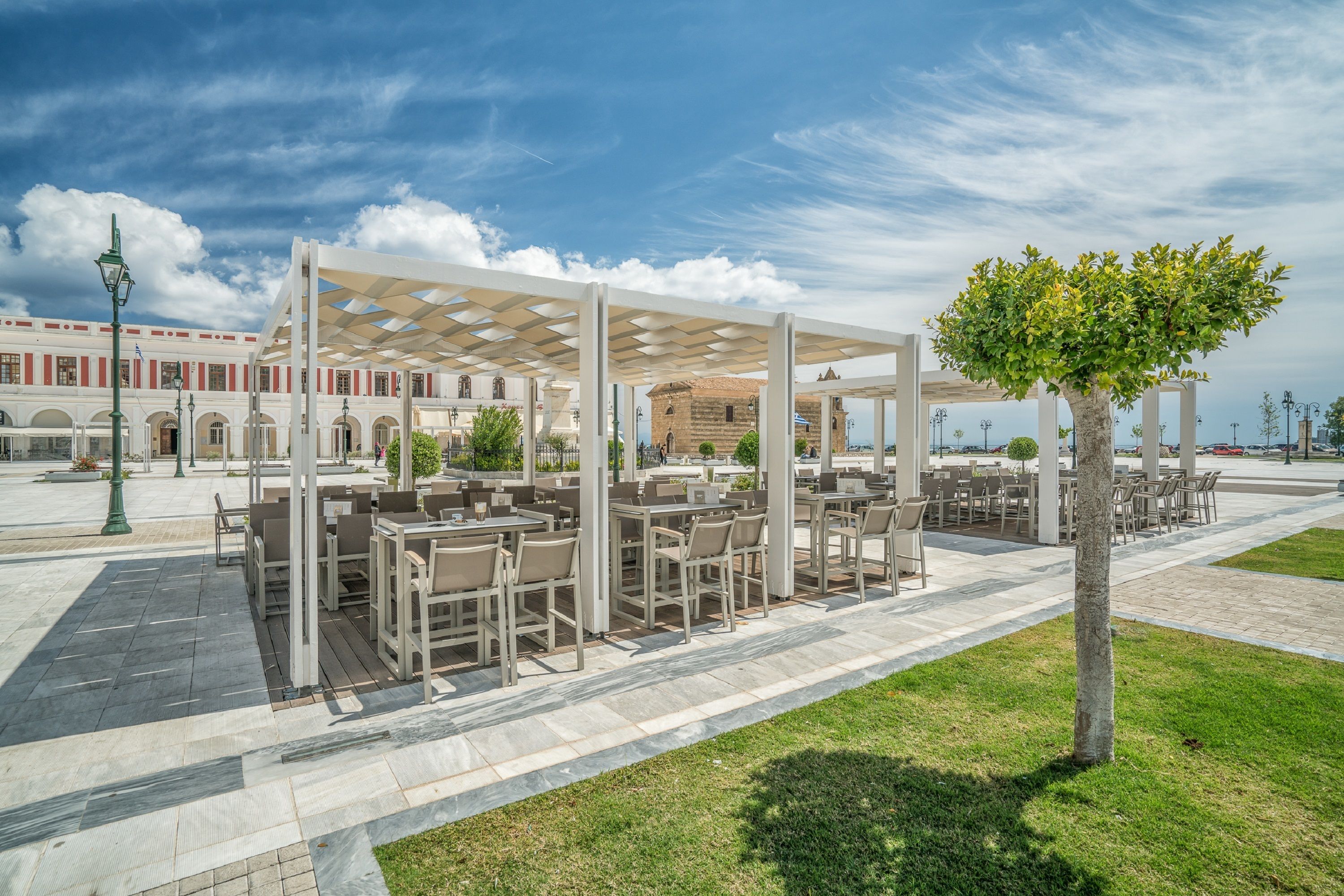 An outdoor café with tables and chairs under a pergola, set against a backdrop of a historic building and a clear sky.