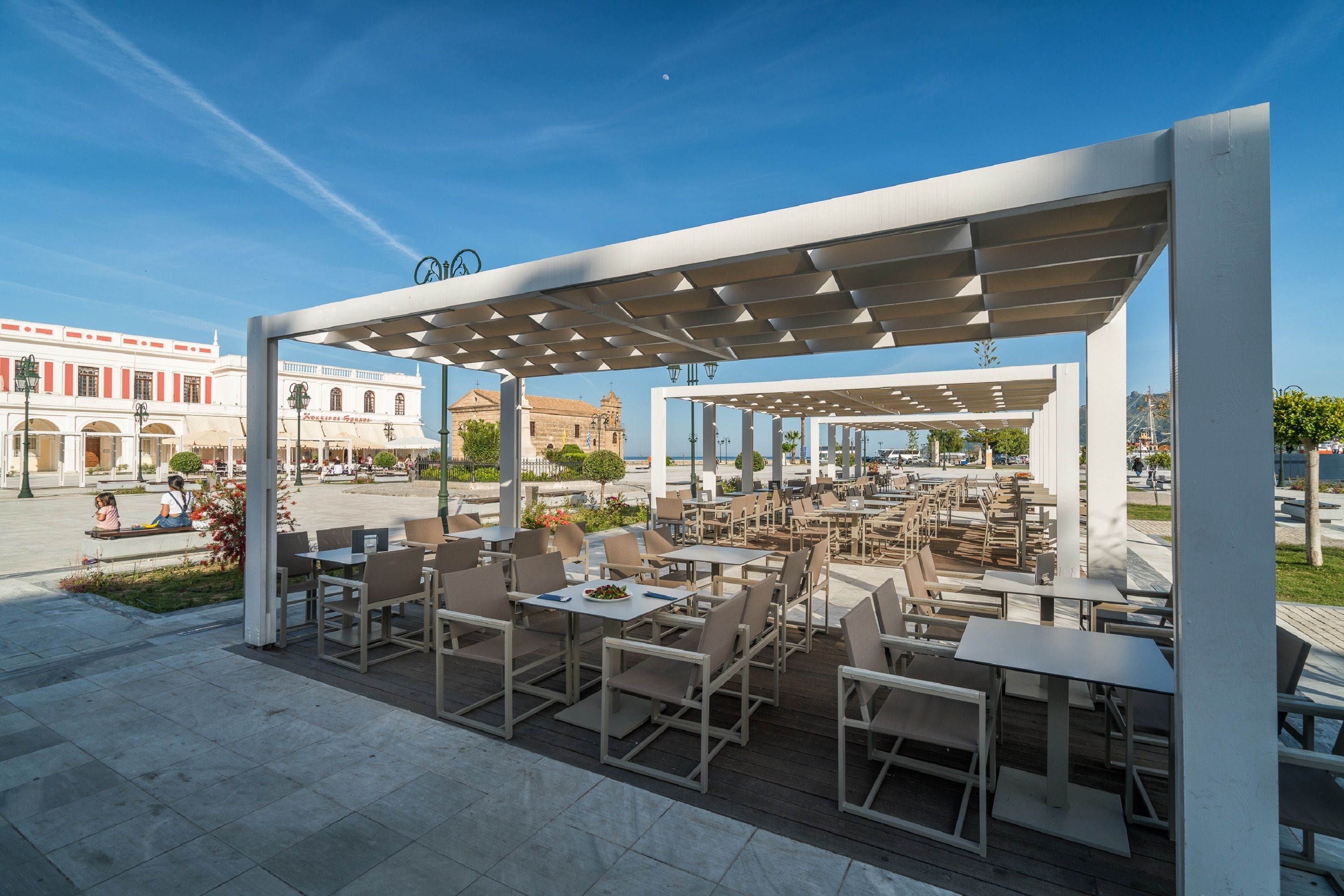 Un café al aire libre con mesas y sillas, una pérgola y vista a un edificio histórico al fondo.