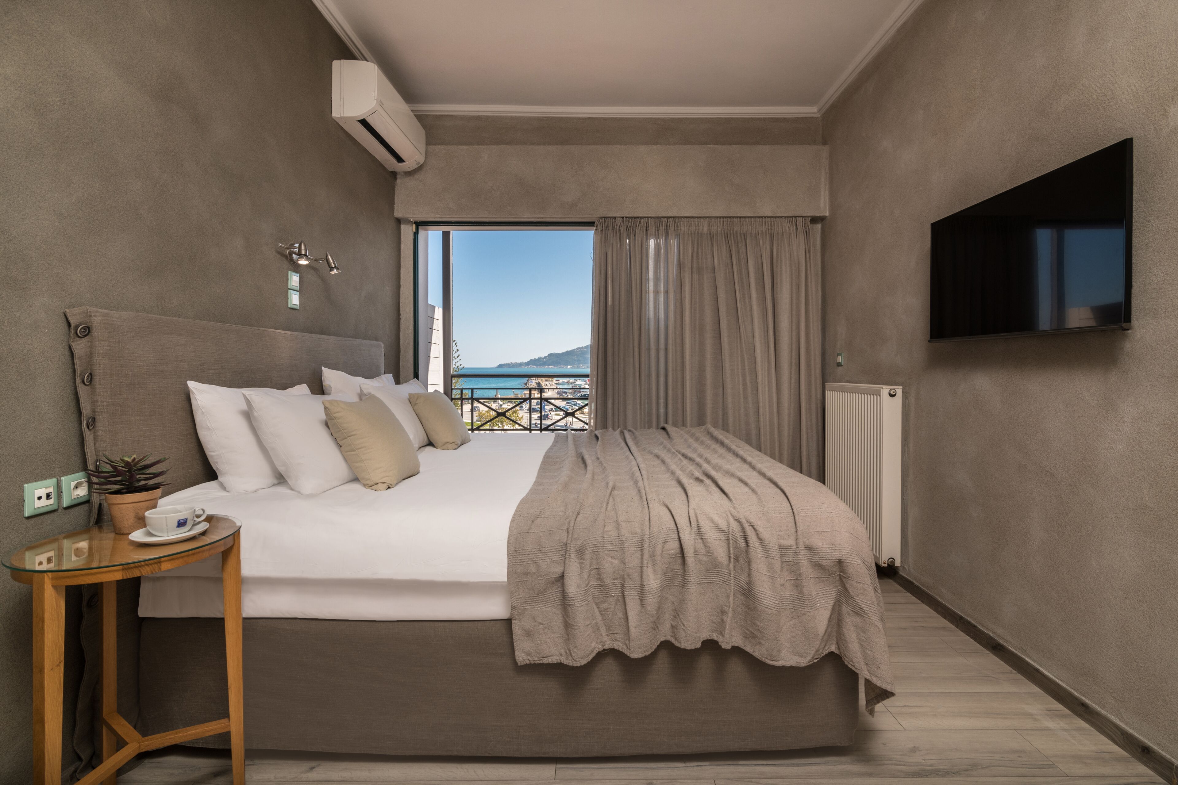 Habitación de hotel moderna con una cama grande, un televisor, una mesita con una taza y un platillo, y vista al mar a través de una ventana.