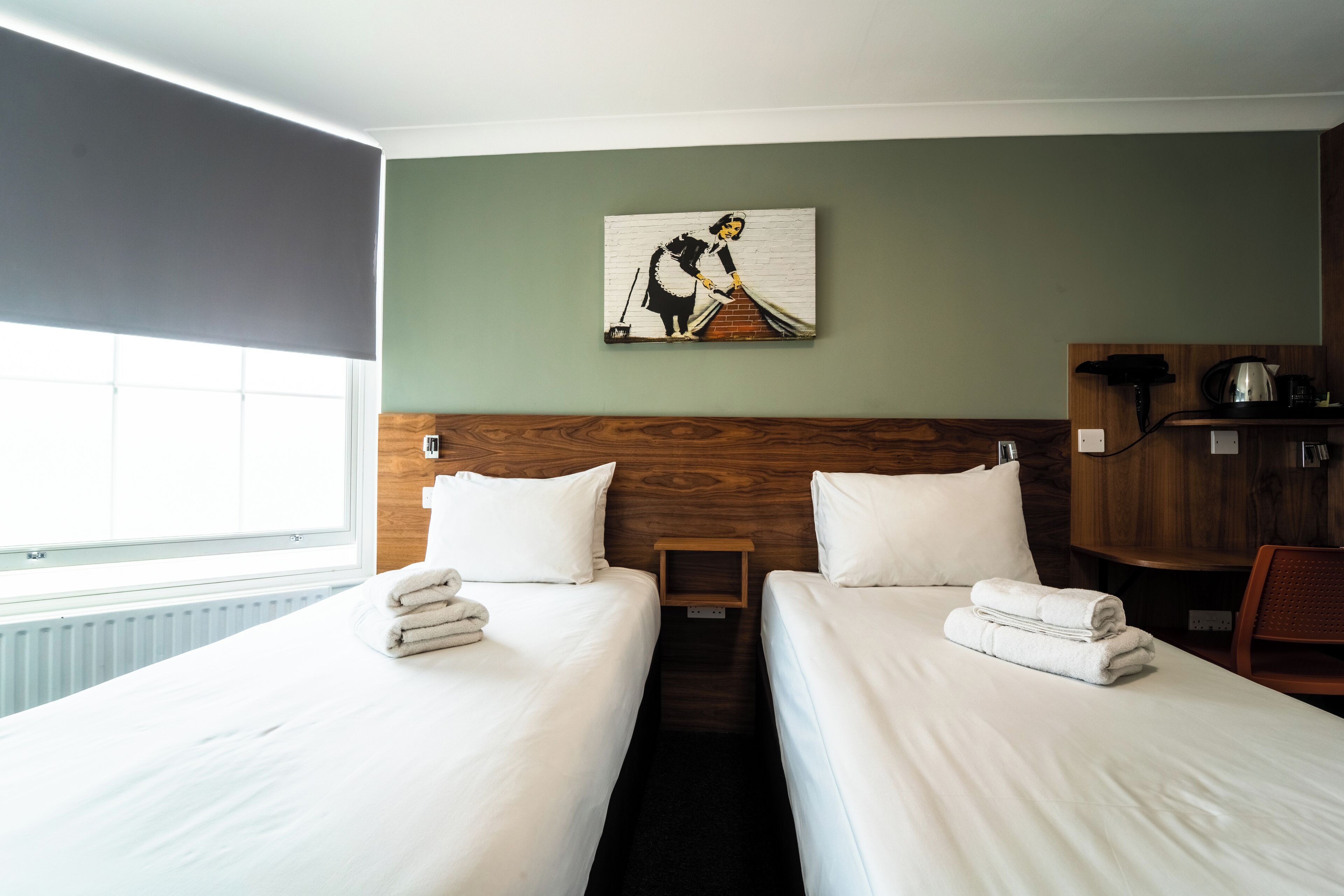 Een hotelkamer met twee bedden, een schilderij aan de muur, een houten hoofdeinde, een tafeltje en een stoel.