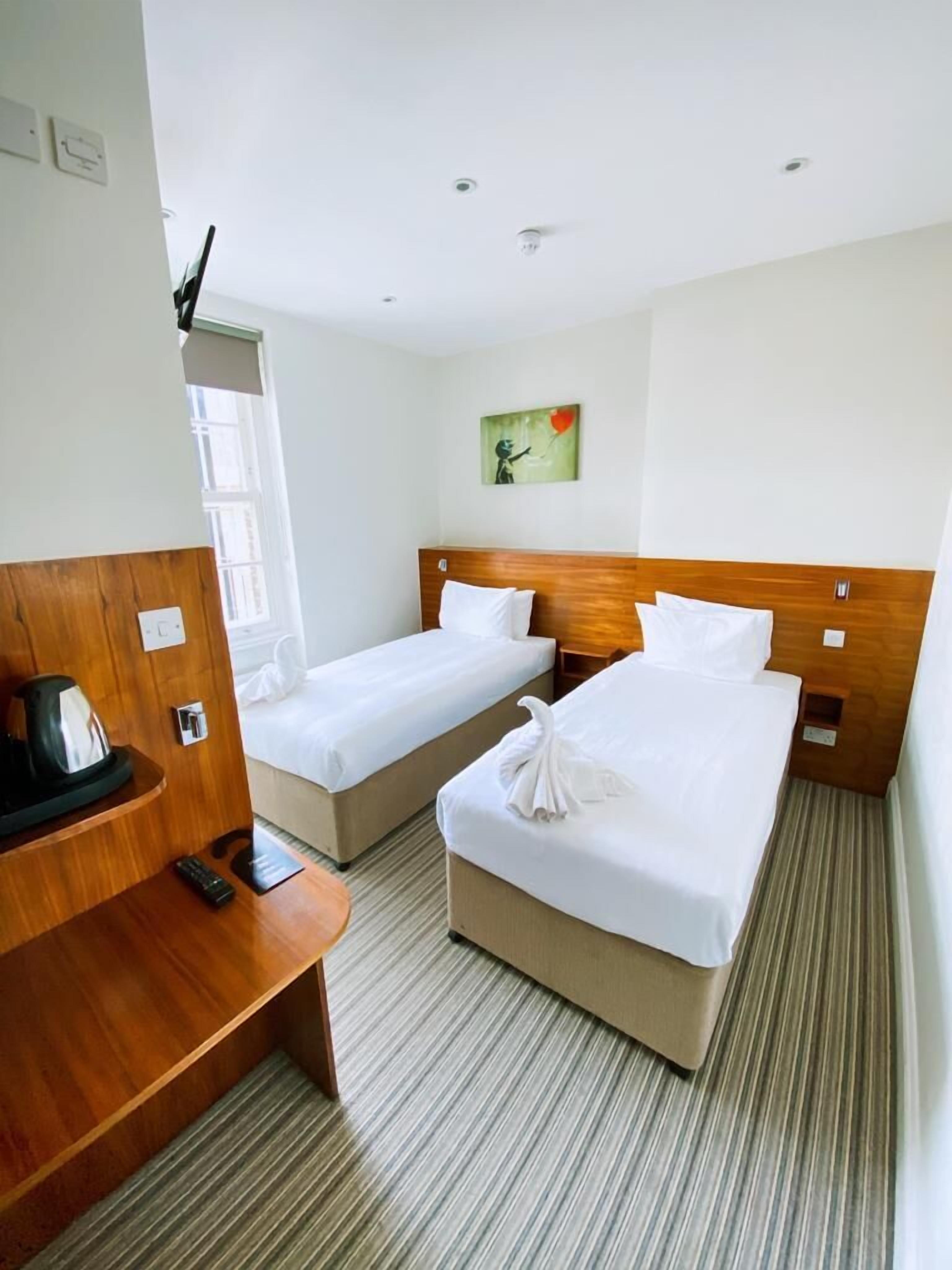 Quarto de hotel com duas camas de solteiro, cabeceira de madeira, uma mesinha com chaleira e um quadro na parede.
