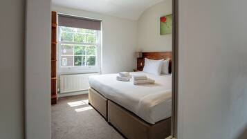 A bedroom with a large window, a bed with pillows and folded towels, a bedside table, and a radiator.