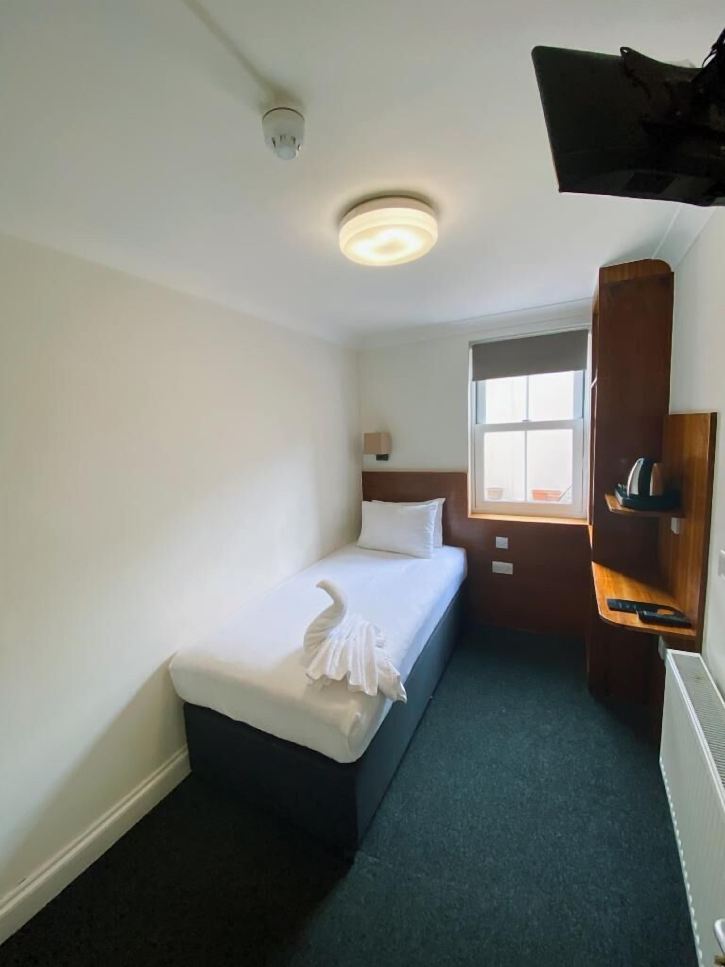 A small, neatly arranged room with a single bed, a window, a radiator, and a shelf with a remote control.