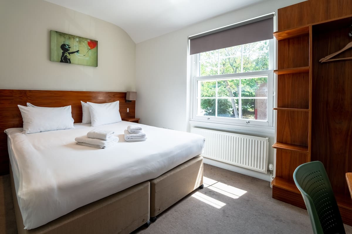 A hotel room with a large bed, a wooden headboard, a window with a view of greenery, and a wardrobe.