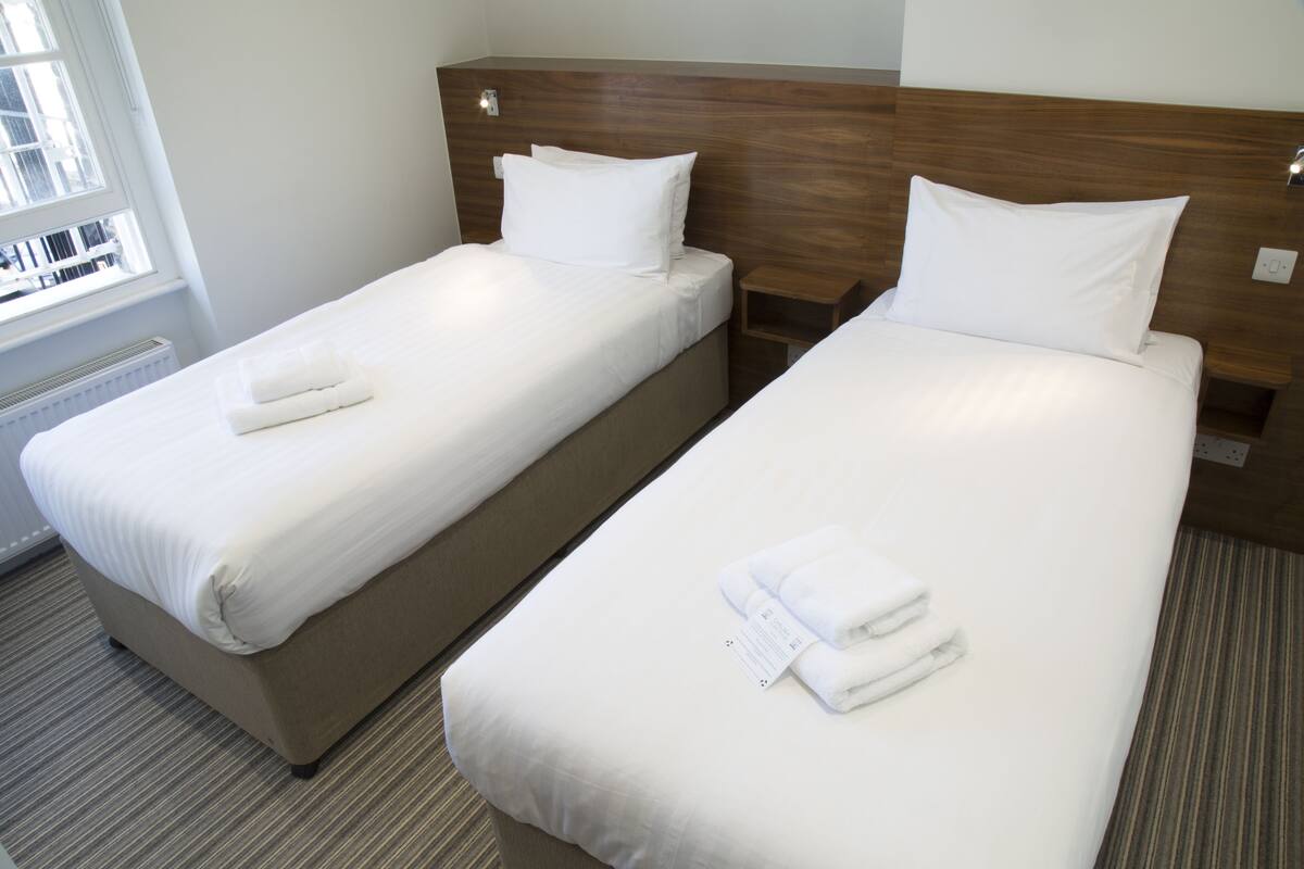 Two single beds with white linens and towels, a wooden headboard, a nightstand, and a window with blinds.