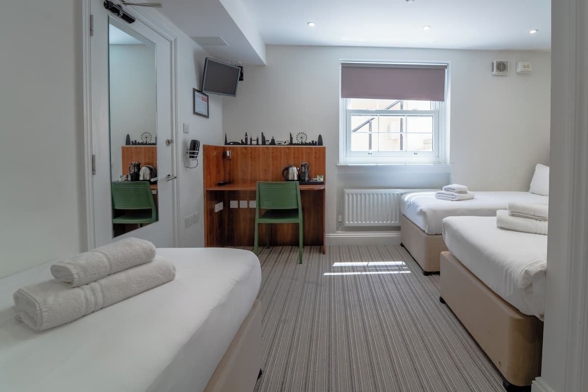 A hotel room with two single beds, a desk with a chair, a radiator, a window with a view, and a television on the wall.