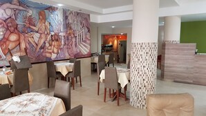 A restaurant with tables set for dining, a large mural on the wall, and a reception area with a green wall. - Hotel Catedral (Mar del Plata)