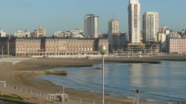 A coastal cityscape with a prominent high-rise building, a waterfront promenade, and a historic building with a row of arches. - Hotel Catedral (Mar del Plata)