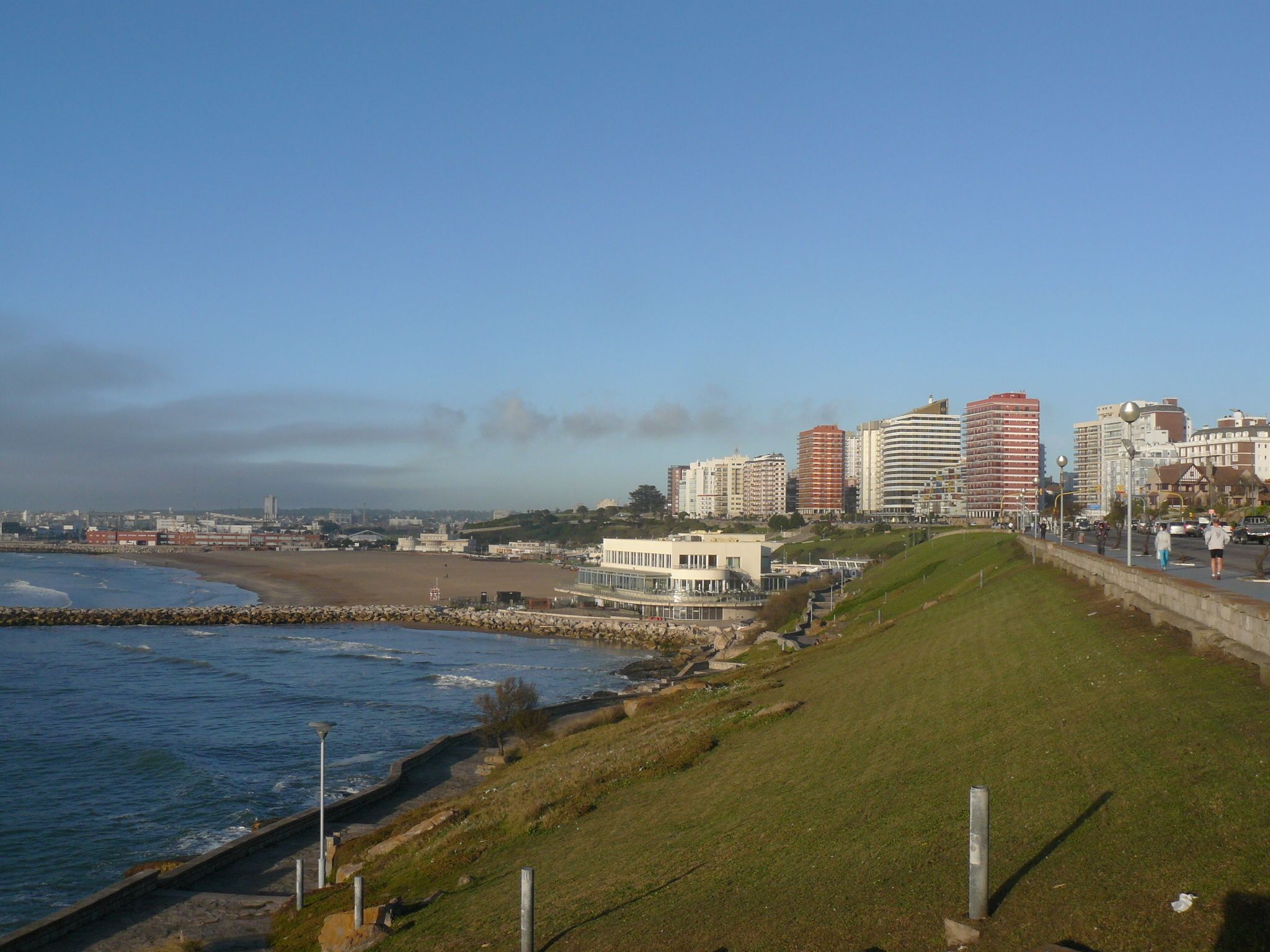 A coastal cityscape with a beach, a grassy embankment, and a row of modern buildings.