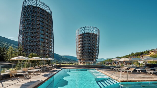 Una piscina con sillas de descanso y sombrillas, rodeada por dos edificios cilíndricos con fachada de celosía.