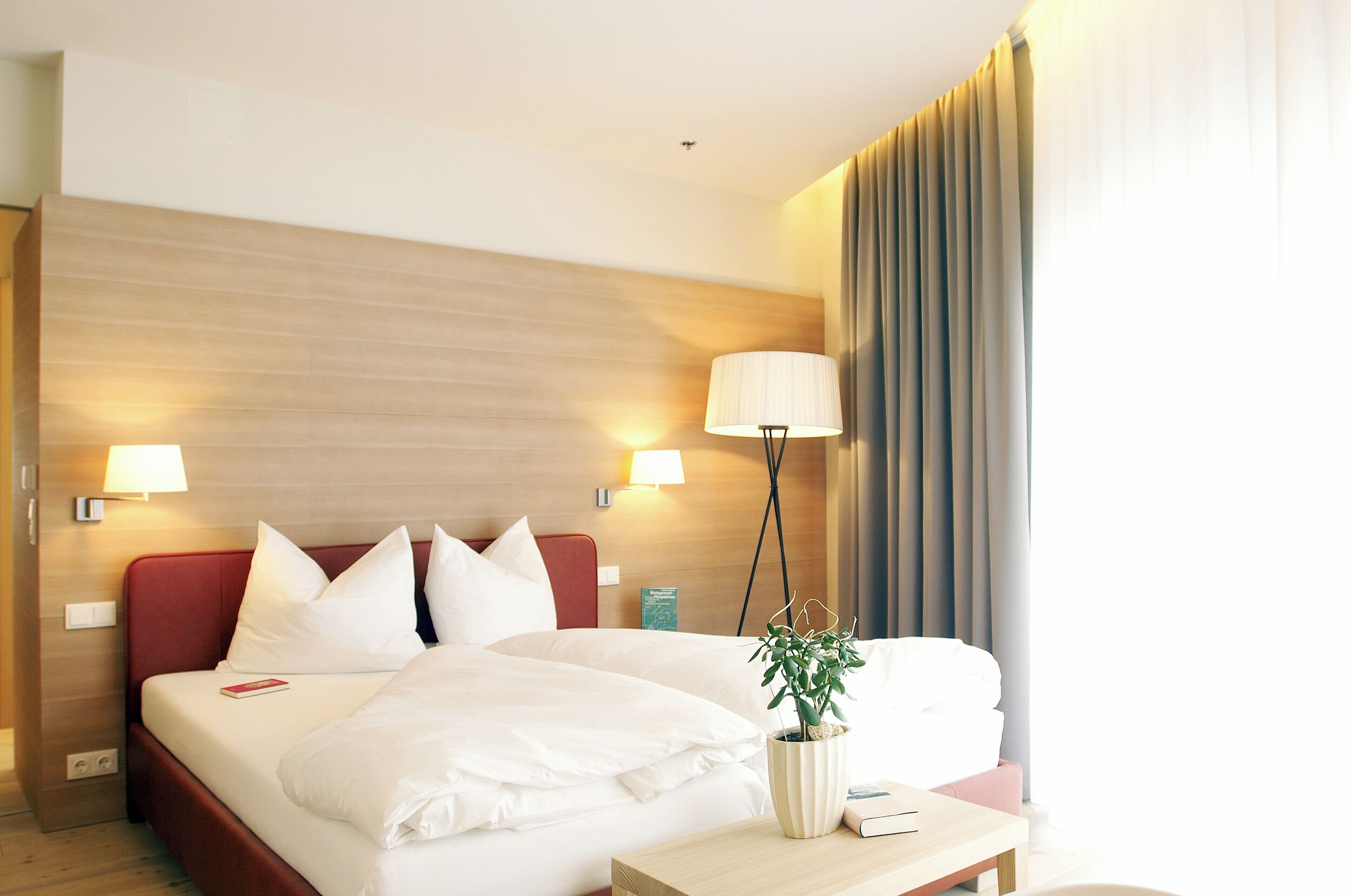 Quarto de hotel com cama, luminárias de cabeceira, mesa de cabeceira e janela com cortinas.