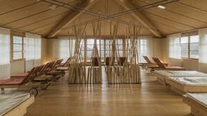 Una habitación amplia con vigas de madera, techo inclinado y ventanas grandes. Hay camas de madera y sillones reclinables dispuestos a lo largo de las paredes.