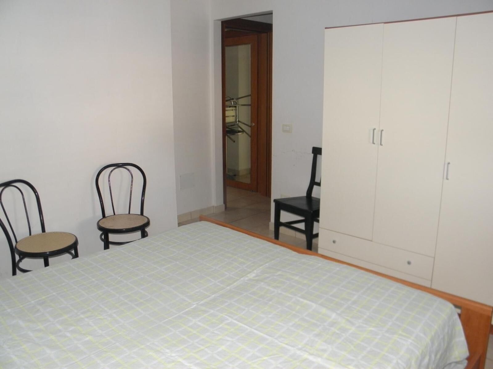 A bedroom with a bed, two chairs, a wardrobe, and a door leading to another room.