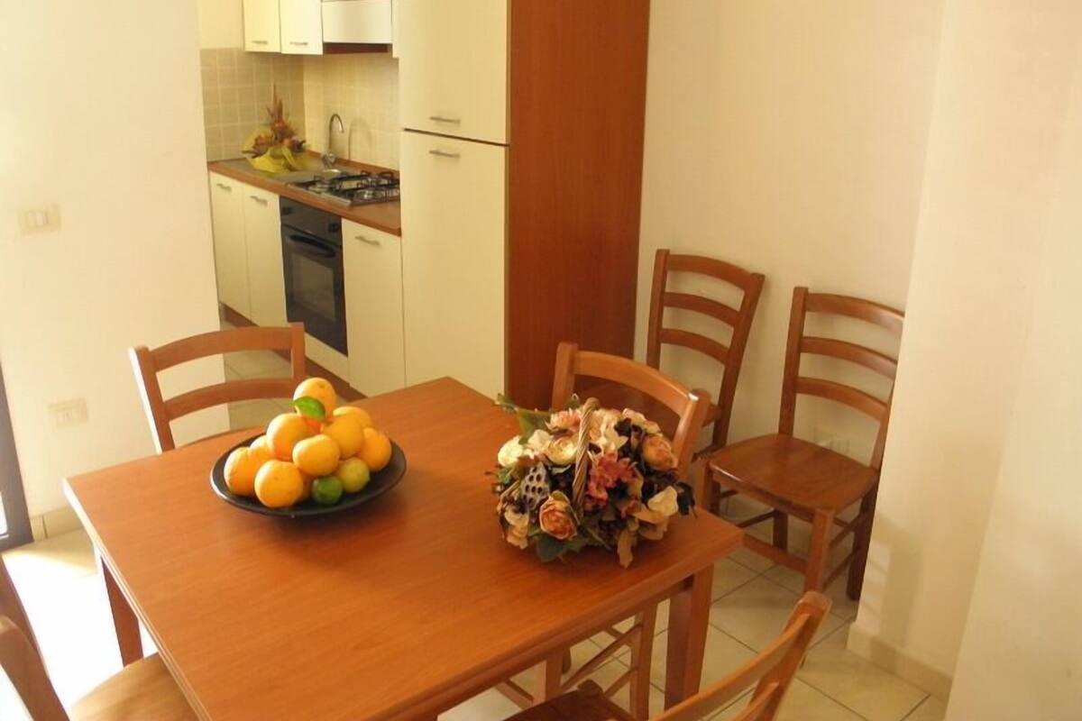 A dining area with a wooden table, four chairs, a plate of fruit, and a bouquet of flowers.
