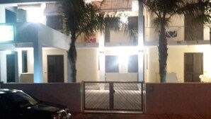 Um edifício de dois andares com entrada fechada por portão, palmeiras e um carro estacionado.